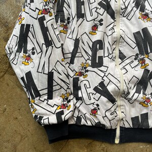 May include: White zip-up jacket featuring a repeating pattern of black letters and Mickey Mouse cartoon characters in red, yellow, and black. The jacket has a black ribbed hem and cuffs, and a white zipper.
