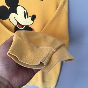 May include: Yellow sweatshirt featuring a smiling Mickey Mouse graphic. The cuff of the long sleeve is visible, showcasing the ribbed knit fabric. The sweatshirt is a solid yellow color.