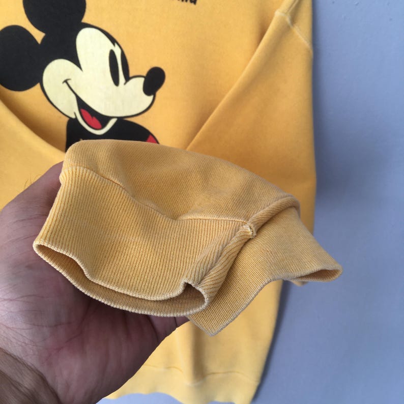 May include: Yellow sweatshirt featuring a smiling Mickey Mouse graphic. The cuff of the long sleeve is visible, showcasing the ribbed knit fabric. The sweatshirt is a solid yellow color.