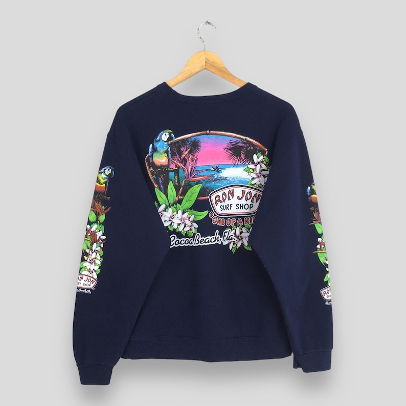 Puede incluir: Sudadera azul marino con dise&ntilde;o tropical, con un loro, palmeras y el texto "Ron Jon Surf Shop Cocoa Beach, Fla." Las mangas tienen dise&ntilde;os florales y de loros adicionales. La sudadera est&aacute; colgada en una percha de madera.