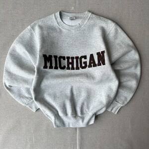 May include: A light grey crewneck sweatshirt featuring the word MICHIGAN printed in large, dark, block-style letters across the chest. The garment has long sleeves, a ribbed collar, and ribbed cuffs, constructed from a heathered fabric material.
