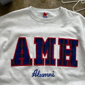 May include: A white crewneck sweatshirt with a speckled texture featuring the large block letters AMH in blue and red plaid fabric with red trim. Below the letters, the word Alumni is embroidered in blue script. A Rugged Wear label is at the neck.