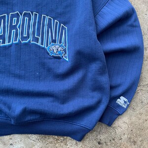 May include: A navy blue long-sleeve sweatshirt with a vertical ribbed texture. The front features the word CAROLINA in large white and light blue letters, with a Tar Heels ram logo. A white Starter logo is embroidered on the left sleeve cuff.