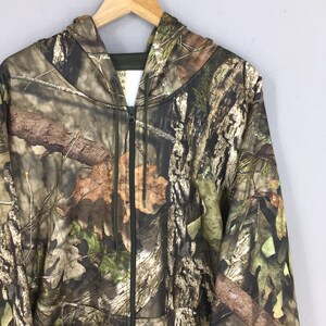 K&ouml;nnte beinhalten: Ein Camouflage-Kapuzenpullover mit Rei&szlig;verschluss und Kapuze, der an einem Holzb&uuml;gel h&auml;ngt. Die Jacke hat ein Wald-Camouflage-Muster in Braun-, Gr&uuml;n- und Beiget&ouml;nen, das eine Waldumgebung nachahmt. Der Rei&szlig;verschluss ist schwarz, und die Jacke hat ein Etikett mit Text.