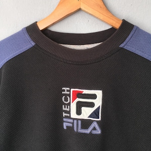 May include: Black FILA sweatshirt with blue shoulder accents. The sweatshirt features a textured pattern and a FILA logo patch with red, white, and blue details. The text "TITECH" and "FILA" are also visible.