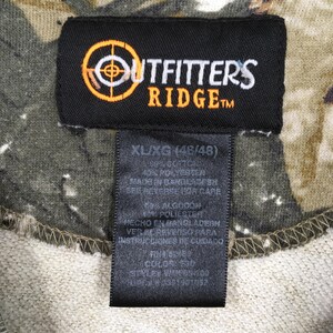 May include: Close-up of a black tag with the "Outfitters Ridge" logo in orange and white. The tag is attached to a camouflage-patterned fabric. The tag also includes size information: XL/XG (46/48).