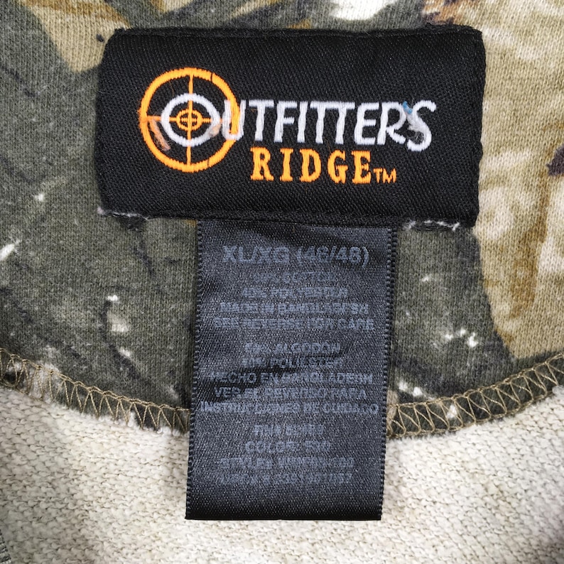 May include: Close-up of a black tag with the "Outfitters Ridge" logo in orange and white. The tag is attached to a camouflage-patterned fabric. The tag also includes size information: XL/XG (46/48).