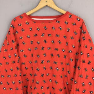 May include: Red long-sleeved sweatshirt with a crew neck and a repeating pattern of a cartoon mouse character. The collar and cuffs are red. The sweatshirt is hanging on a wooden hanger.