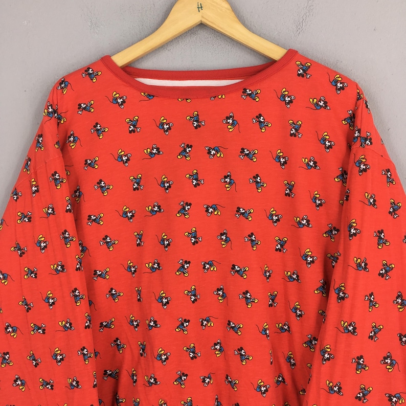 May include: Red long-sleeved sweatshirt with a crew neck and a repeating pattern of a cartoon mouse character. The collar and cuffs are red. The sweatshirt is hanging on a wooden hanger.