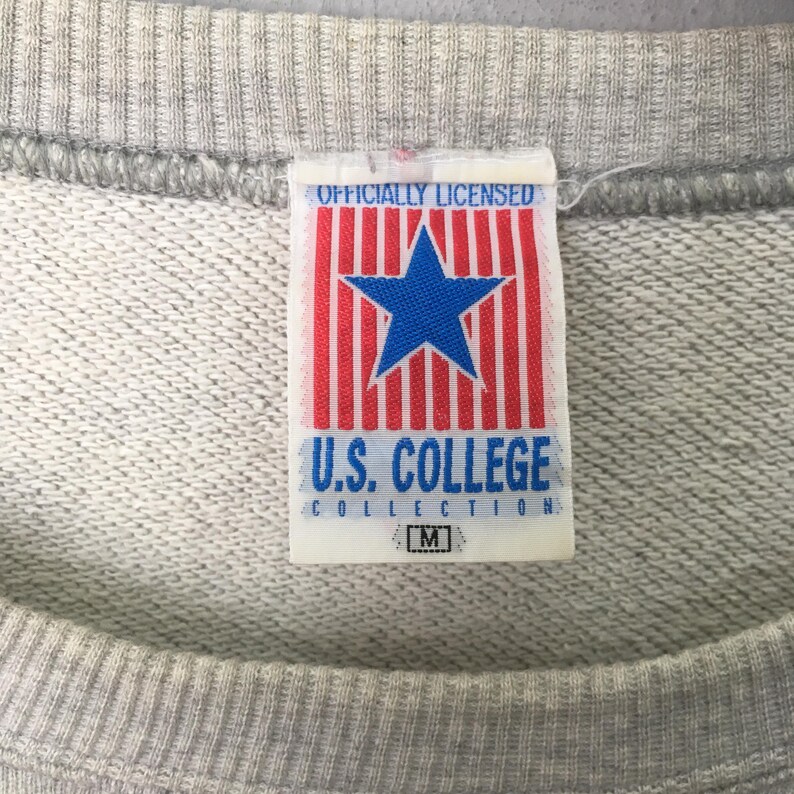 Puede incluir: Una sudadera gris claro con una etiqueta de tela blanca en el cuello. La etiqueta muestra el texto "OFFICIALLY LICENSED" sobre una estrella azul centrada en rayas verticales rojas, con "U.S. COLLEGE COLLECTION" y la talla "M" debajo.