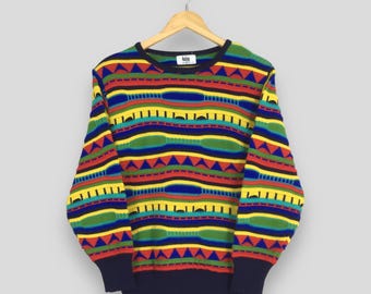 Puede incluir: Un suéter colorido con un patrón geométrico. El suéter presenta rayas horizontales en rojo, amarillo, verde y azul, con detalles en negro. Los puños y el cuello son de color azul marino. El suéter está colgado de una percha de madera.