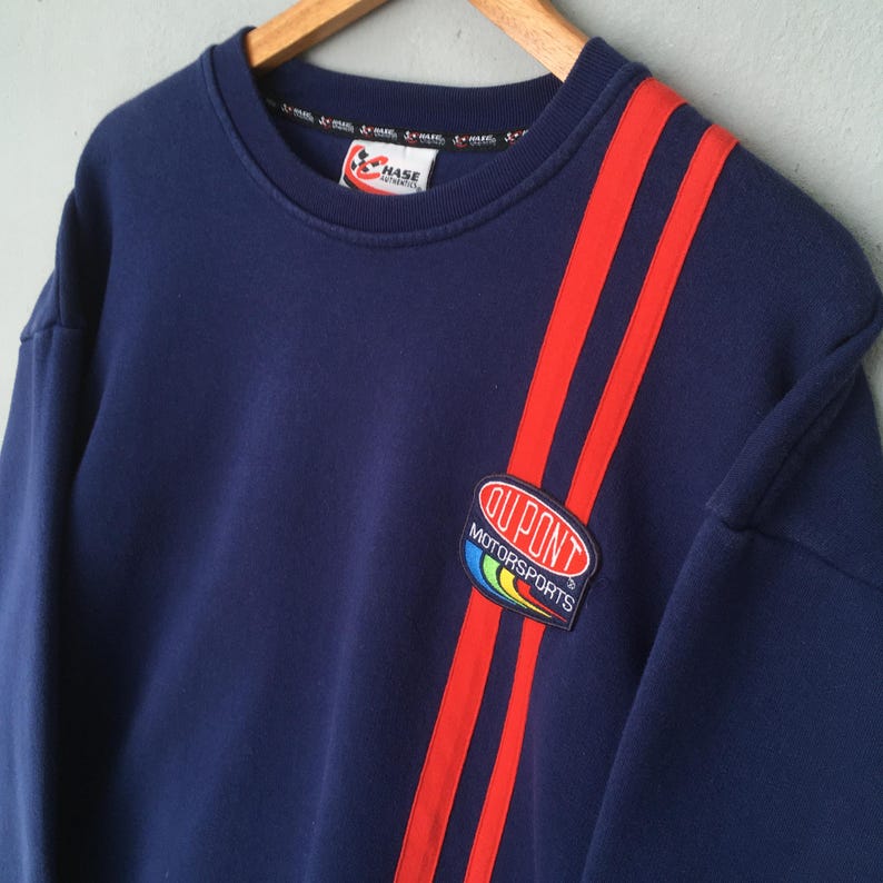 May include: Navy blue sweatshirt with red stripes down the shoulder and a "DuPont Motorsports" patch. The sweatshirt also features a small "Hase" logo on the collar. This vintage crewneck is a classic piece of racing apparel.
