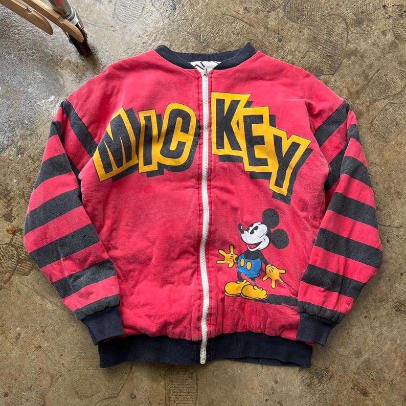 May include: A red zip-up jacket with the word "MICKEY" in yellow block letters across the chest. The jacket features black and gray striped sleeves and a cartoon image of Mickey Mouse. The jacket has a black collar and cuffs.