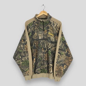 May include: A camouflage-patterned pullover sweatshirt with a quarter-zip neckline. The sweatshirt features a mix of green, brown, and tan colors, with tan-colored sleeves and collar. The Cabela's logo is embroidered in yellow on the chest.