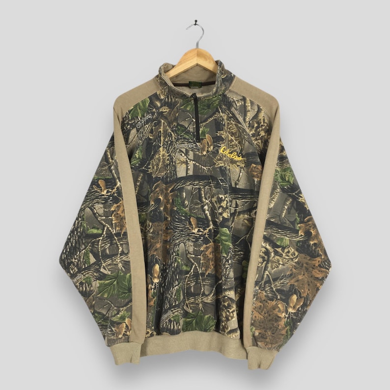May include: A camouflage-patterned pullover sweatshirt with a quarter-zip neckline. The sweatshirt features a mix of green, brown, and tan colors, with tan-colored sleeves and collar. The Cabela's logo is embroidered in yellow on the chest.