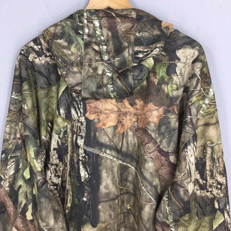 K&ouml;nnte beinhalten: Eine Tarnjacke mit Kapuze in Braun-, Gr&uuml;n- und Beiget&ouml;nen, die sich in eine Waldumgebung einf&uuml;gt. Die Jacke hat eine Kapuze und einen durchgehenden Rei&szlig;verschluss. Das Muster umfasst &Auml;ste, Bl&auml;tter und Rinde.
