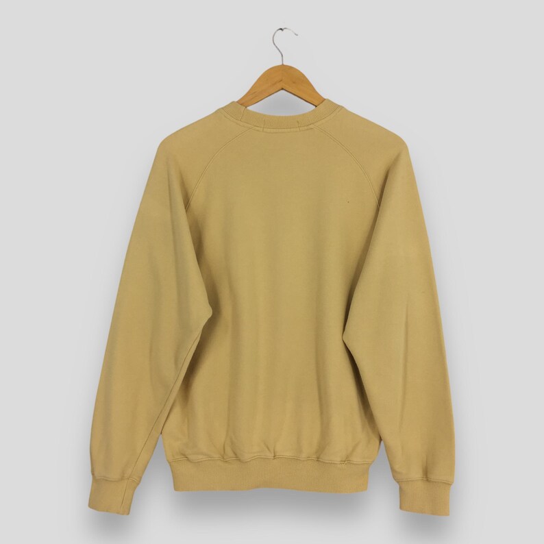 May include: A tan-colored sweatshirt with long sleeves and a crew neck, hanging on a wooden hanger. The raglan sleeves extend from the collar to the underarm. The sweatshirt has ribbed cuffs and a ribbed hem.