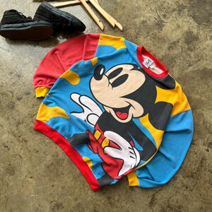K&ouml;nnte beinhalten: Ein Vintage Mickey Mouse Sweatshirt in Blau, Rot und Gelb. Das Sweatshirt zeigt eine gro&szlig;e Grafik von Mickey Mouse. Die &Auml;rmel sind rot mit gelben B&uuml;ndchen und der Halsausschnitt ist rot. Schwarze Turnschuhe sind im Hintergrund zu sehen.