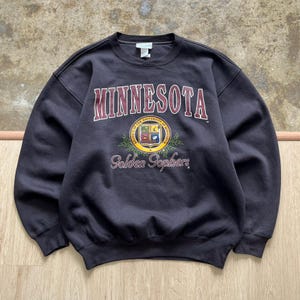 Könnte beinhalten: Ein dunkel anthrazitfarbenes Sweatshirt mit Rundhalsausschnitt, das den Schriftzug MINNESOTA in kastanienbraunen Buchstaben über einem kreisförmigen University of Minnesota Golden Gophers Emblem mit grünen Blättern und dem Text Golden Gophers zeigt.