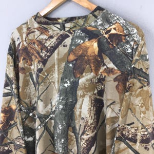 May include: A long-sleeved camouflage shirt in shades of brown, green, and tan, featuring a pattern of leaves and branches. The shirt is hanging on a wooden hanger against a light gray background.