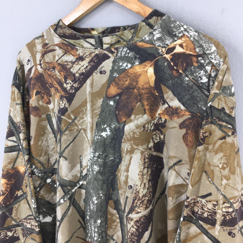 May include: A long-sleeved camouflage shirt in shades of brown, green, and tan, featuring a pattern of leaves and branches. The shirt is hanging on a wooden hanger against a light gray background.