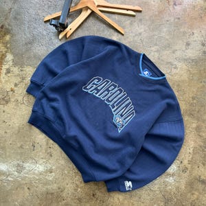 May include: A navy blue long-sleeve sweatshirt featuring the word CAROLINA embroidered in light blue and white lettering across the chest. The garment includes a small ram mascot logo, a ribbed crew neckline with light blue trim, and a Starter logo.