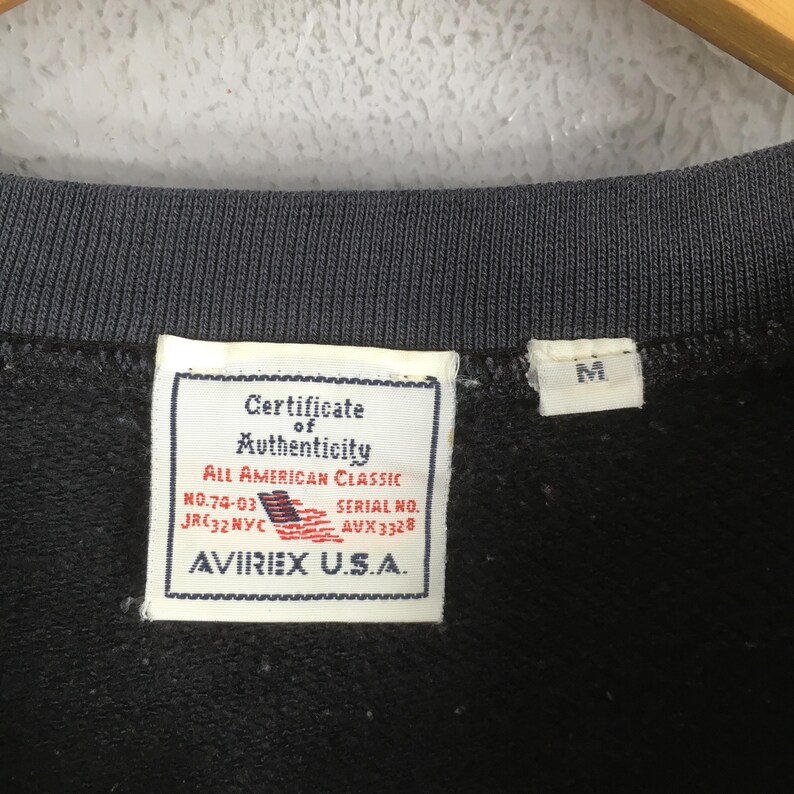 May include: A black garment featuring a white interior tag with the text Certificate of Authenticity, All American Classic, No.74-03, Serial No. AVX3328, and AVIREX U.S.A. A smaller white tag displays the letter M, indicating a medium size.