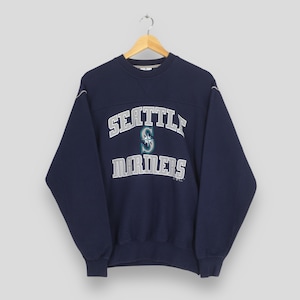May include: Navy blue Seattle Mariners sweatshirt with white and teal lettering. The word "SEATTLE" is arched across the chest, above "MARINERS". The team logo is in the center. The sweatshirt is hanging on a wooden hanger.
