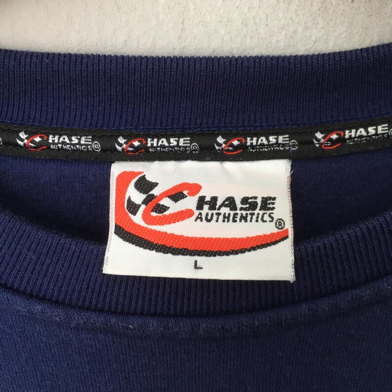 May include: Close-up of a navy blue crew neck shirt. The shirt features a tag with the text "HASE AUTHENTICS" in red and white. The tag also has a size "L" printed on it. The collar has a black band with the text "HASE AUTHENTICS" printed on it.