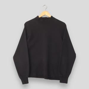 Puede incluir: Una sudadera negra lisa con cuello alto, colgada de una percha de madera. La sudadera tiene mangas largas y puños acanalados. El cuello es alto y ceñido.