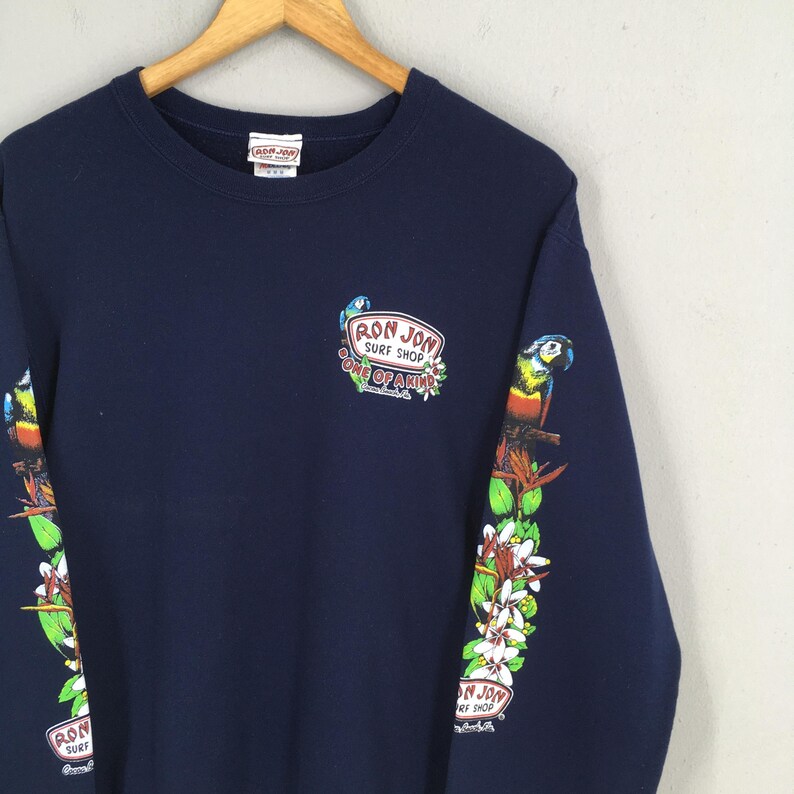 Puede incluir: Sudadera azul marino con el logo "Ron Jon Surf Shop" y gr&aacute;ficos de loros en las mangas. La sudadera tiene cuello redondo y una etiqueta en la parte posterior del cuello.