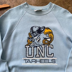 May include: A light blue crewneck sweatshirt featuring a graphic of a ram wearing a hat with the text UNC. Below the ram, the text UNC is printed in large block letters, followed by the word TARHEELS in a smaller font, all in navy and white.