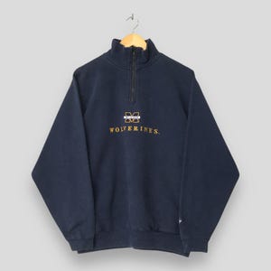 May include: A navy blue quarter-zip sweatshirt featuring a high collar with a zip closure. The chest displays an embroidered Michigan logo in white and yellow, with the word WOLVERINES stitched in yellow lettering below the logo.