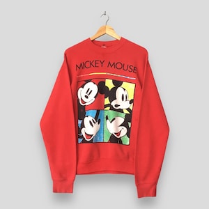 May include: Red crewneck sweatshirt featuring a four-panel graphic of Mickey Mouse with different facial expressions. The words "MICKEY MOUSE" are printed across the top in black. The sweatshirt is hanging on a wooden hanger.