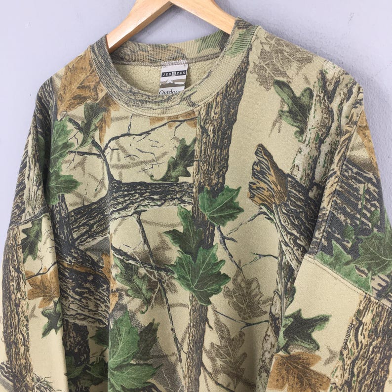 May include: A camouflage-patterned sweatshirt with a crew neck and long sleeves. The design features a mix of brown, green, and tan colors, mimicking a forest environment. The tag reads "Outdoors".