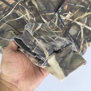 May include: Close-up of a camouflage-patterned long-sleeve shirt. The fabric features a blend of brown, green, and tan hues, mimicking a natural woodland environment. The cuff of the sleeve is visible, showcasing the ribbed texture and color variations.