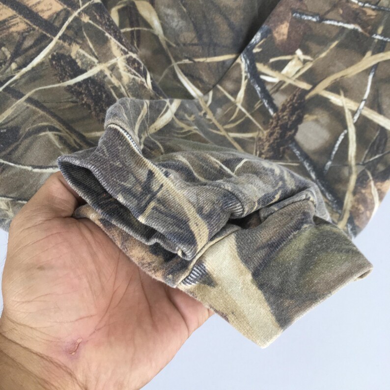 May include: Close-up of a camouflage-patterned long-sleeve shirt. The fabric features a blend of brown, green, and tan hues, mimicking a natural woodland environment. The cuff of the sleeve is visible, showcasing the ribbed texture and color variations.