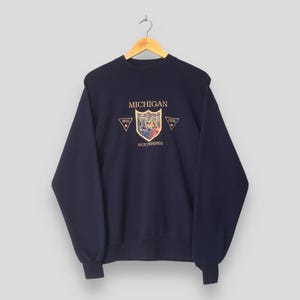 May include: A navy blue long-sleeve sweatshirt featuring an embroidered crest on the chest. The crest displays the letter M, the text MICHIGAN WOLVERINES, and the words VERITAS and SCIENTIA inside small triangles on either side of the shield.