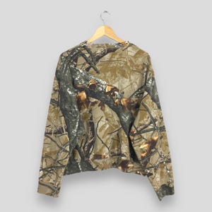 May include: A long-sleeved camouflage sweatshirt hangs on a wooden hanger. The pattern features shades of brown, green, and gray, mimicking a forest environment. The sweatshirt is made of a soft material and has a crew neck.