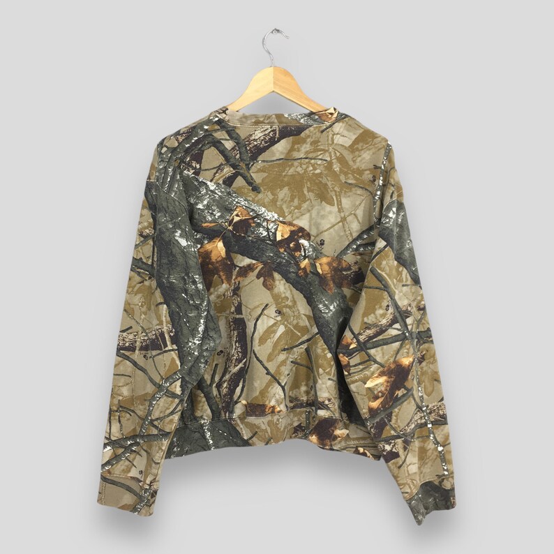 May include: A long-sleeved camouflage sweatshirt hangs on a wooden hanger. The pattern features shades of brown, green, and gray, mimicking a forest environment. The sweatshirt is made of a soft material and has a crew neck.
