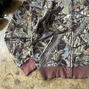 K&ouml;nnte beinhalten: Ein Kapuzenpullover mit Rei&szlig;verschluss und Tarnmuster in Gr&uuml;n-, Braun- und Graut&ouml;nen. Die B&uuml;ndchen und der Bund sind einfarbig braun. Der Hoodie hat einen Frontrei&szlig;verschluss und zwei Taschen.