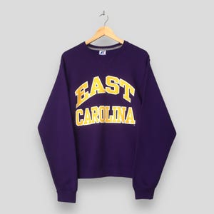 以下が含まれることがあります： 胸元に黄色いブロック体でEAST CAROLINAの文字がプリントされた、紫色の長袖クルーネックトレーナー。襟元、袖口、裾はリブ編みになっており、ネックラインの下にはV字型のステッチが施されています。