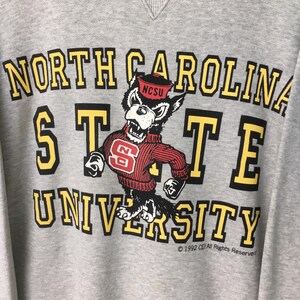Puede incluir: Una sudadera gris jaspeado con el texto NORTH CAROLINA STATE UNIVERSITY en letras de bloque amarillas y negras. Un gr&aacute;fico de una mascota de lobo con un su&eacute;ter rojo con un logotipo NC y una gorra roja est&aacute; centrado entre las palabras.