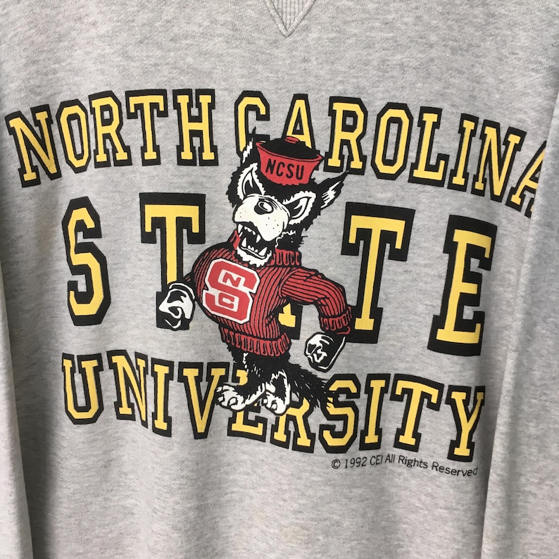 Puede incluir: Una sudadera gris jaspeado con el texto NORTH CAROLINA STATE UNIVERSITY en letras de bloque amarillas y negras. Un gr&aacute;fico de una mascota de lobo con un su&eacute;ter rojo con un logotipo NC y una gorra roja est&aacute; centrado entre las palabras.