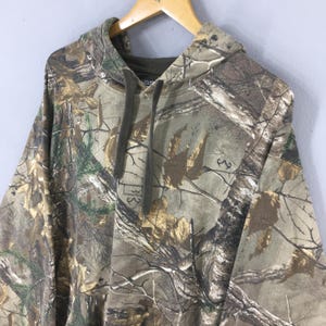 Op de afbeelding: Een hoodie met camouflagepatroon, weergegeven op een houten hanger. Het ontwerp heeft een mix van bruine, groene en bruine tinten, die een bosrijke omgeving nabootsen. De hoodie heeft een relaxte pasvorm en lange mouwen.