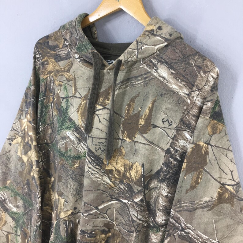 Op de afbeelding: Een hoodie met camouflagepatroon, weergegeven op een houten hanger. Het ontwerp heeft een mix van bruine, groene en bruine tinten, die een bosrijke omgeving nabootsen. De hoodie heeft een relaxte pasvorm en lange mouwen.