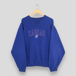 May include: A blue sweatshirt with the word "KANSAS" embroidered in red, with a small graphic below. The sweatshirt is hanging on a wooden hanger. The garment has a crew neck and long sleeves.