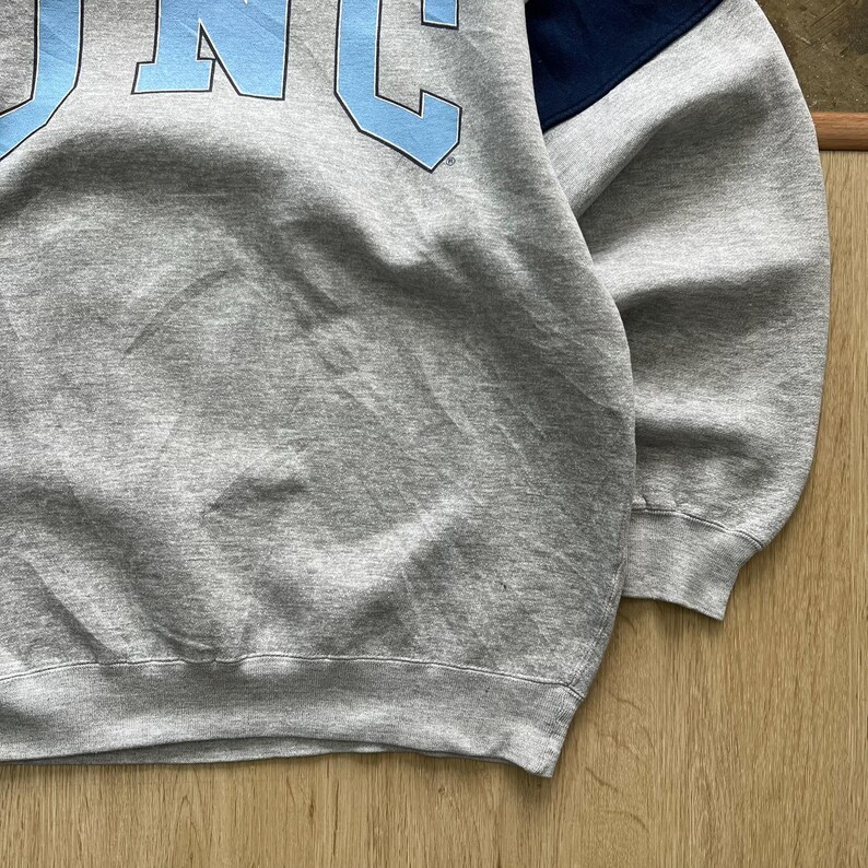 Puede incluir: Una sudadera gris jaspeado con las letras "UNC" en azul claro en el pecho. La parte superior derecha del hombro es azul marino. La sudadera tiene cuello redondo y mangas largas. La tela parece ser un material suave y c&oacute;modo.