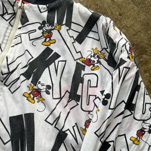 May include: White zip-up jacket featuring a repeating pattern of Mickey Mouse and the word "Mickey" in black. The jacket has a black collar and a red zipper detail. Mickey Mouse is depicted in his classic red shorts, yellow shoes, and white gloves.