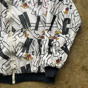 May include: White zip-up jacket featuring a repeating pattern of black letters and Mickey Mouse illustrations in red, yellow, and black. The jacket has a black ribbed collar and cuffs, and a white zipper.